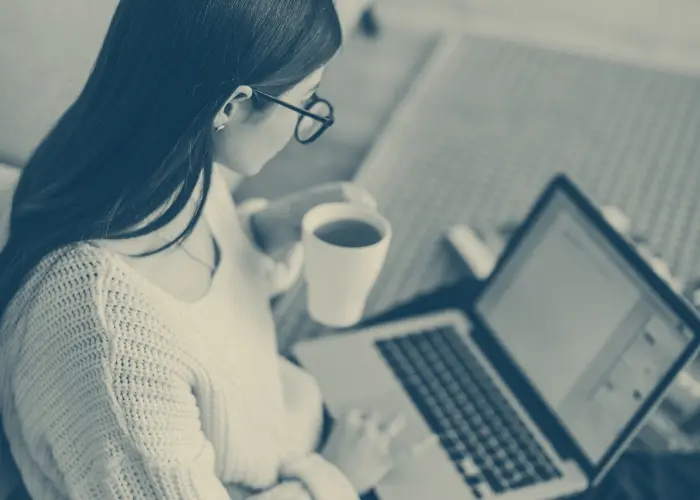 Woman with glasses holding a cup and looking at a laptop.