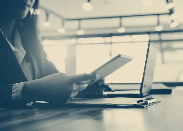 An image of a person holding a tablet at a table with a laptop and papers.